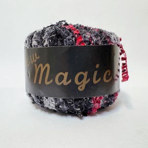 May include: A ball of New Magic yarn, featuring a mix of black, red, and silver threads. The yarn is wrapped with a black label that has the words "New Magic" in gold lettering. The yarn has a textured appearance.