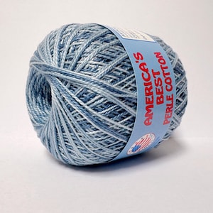 May include: A ball of blue and white variegated perle cotton yarn. The yarn is wrapped in a ball shape with a blue label that reads "America's Best Perle Cotton". The yarn is a mix of light and dark blue shades.