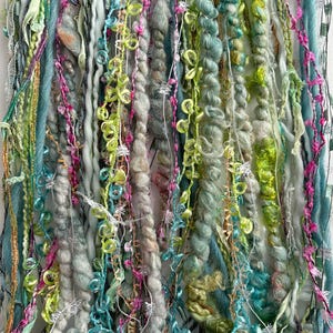 May include: A collection of hanging fiber art strands featuring a variety of textures and colors including shades of teal, lime green, mint, white, and magenta. The strands consist of twisted yarn, fuzzy wool, metallic threads, and looped ribbons.