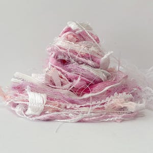 May include: A pile of pink, white, and silver craft yarn and ribbon. The yarn is a mix of textures, including fluffy, smooth, and metallic threads. A white lace ribbon is visible on the right side. The overall effect is soft and delicate.
