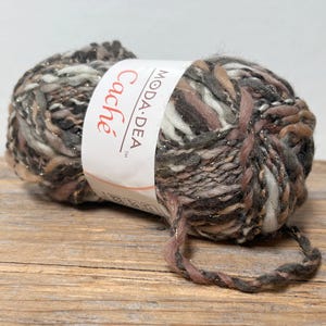 May include: A ball of Cache yarn by Moda Dea, in shades of gray, brown, and pink, with metallic gold threads. The yarn is resting on a wooden surface.