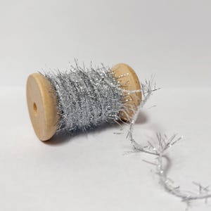 May include: A wooden spool wrapped with silver tinsel. The tinsel has a metallic sheen and is loosely wound around the spool. The spool is made of light-colored wood and has a hole in the center.