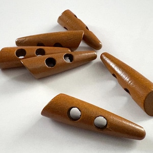 May include: A collection of six wooden toggle buttons in a warm brown hue. Each button is cylindrical with a rounded end and two or three holes for sewing. The buttons are ideal for coats, sweaters, and other garments.