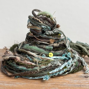 May include: A coiled bundle of assorted decorative ribbons and yarns in shades of sage green, dark brown, and metallic silver. The collection features varied textures, including fuzzy wool, braided cord, and a single bright yellow sequin accent.