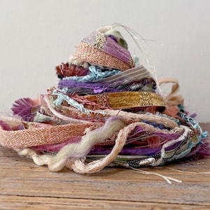 May include: A pile of colorful craft ribbon and yarn in shades of pink, purple, gold, and teal. The ribbons are various textures, including woven, beaded, and fringed. The pile is sitting on a wooden surface.