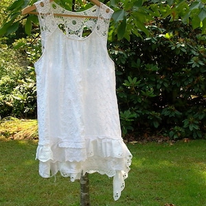 May include: White crochet lace sleeveless top with a ruffled hem and a scalloped neckline. The top is hanging on a hanger against a green background.