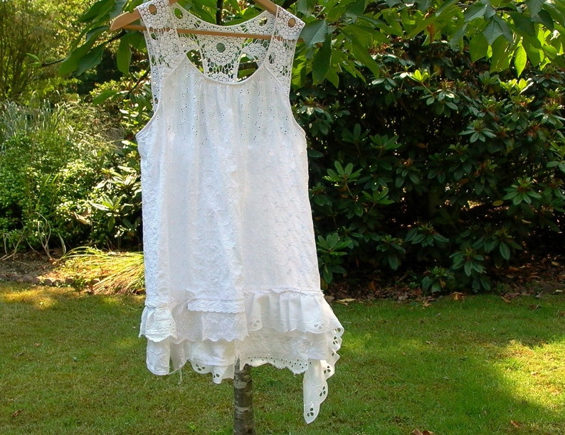 May include: White crochet lace sleeveless top with a ruffled hem and a scalloped neckline. The top is hanging on a hanger against a green background.