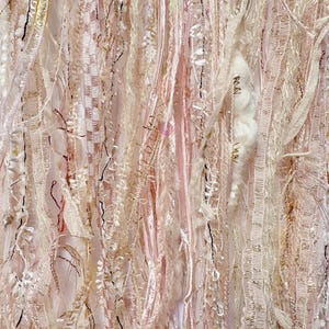 May include: Close-up of a textile artwork featuring a layered composition of ribbons, threads, and fabrics in shades of pink, cream, and beige. The textures include satin, lace, and woven elements, with accents of black thread and metallic gold.