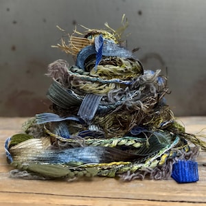 May include: A tangled pile of assorted ribbons and yarns in shades of olive green, slate blue, metallic gold, and charcoal gray. The collection features various textures including frayed edges, braided strands, and smooth satin-like ribbons.