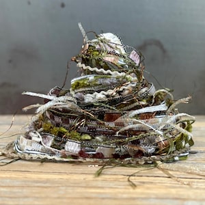 May include: A coiled bundle of decorative ribbons and textured yarns in shades of olive green, brown, white, and metallic silver. The assortment includes various widths, frayed edges, and shimmering threads layered in a dense, circular stack.