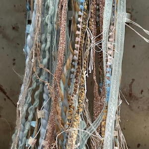 May include: Close-up of various strands of decorative yarn and ribbon in shades of blue, beige, and copper. The textures include braided, woven, and fringed elements, creating a layered and textured appearance. Ideal for craft projects.
