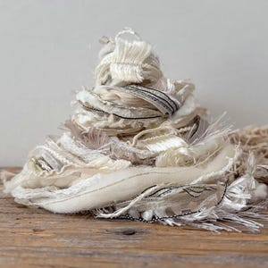 May include: A pile of off-white and cream-colored fabric scraps with frayed edges and black stitching. The fabric is arranged in a conical shape on a wooden surface, suggesting a craft supply or decorative element.