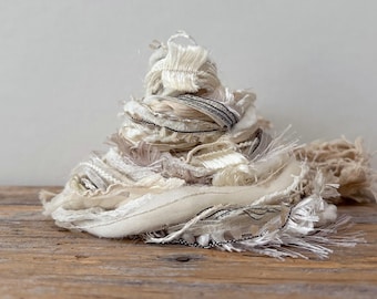 May include: A pile of off-white and cream-colored fabric scraps with frayed edges and black stitching. The fabric is arranged in a conical shape on a wooden surface, suggesting a craft supply or decorative element.