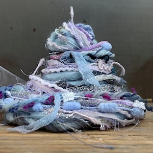 May include: A decorative bundle of assorted textile fibers and ribbons in shades of light blue, lavender, and silver. The collection includes textured yarns, frayed fabric strips, metallic threads, and small pom-poms, all coiled into a layered heap.