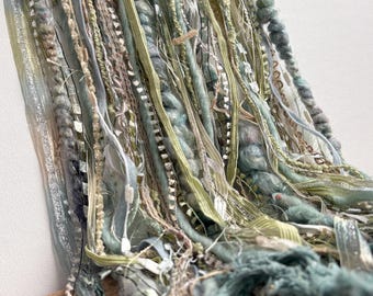 May include: A collection of hanging fiber art strands in shades of sage green, olive, and cream. The composition features a variety of textures including thick wool yarn, shimmering metallic ribbons, frayed fabric strips, and delicate lace trim.