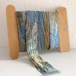 May include: A spool of multi-colored ribbon with a blue, green, and brown ombre effect. The ribbon is wrapped around a cardboard spool with a hole in the center.