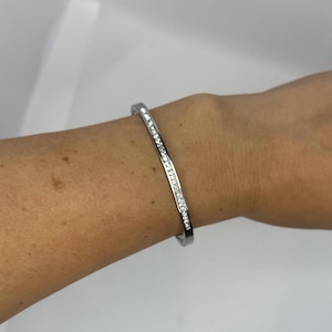 May include: A silver bracelet with a line of small, clear gemstones. The bracelet is curved and worn on a wrist. The background is plain white. The gemstones are set along the top of the bracelet.