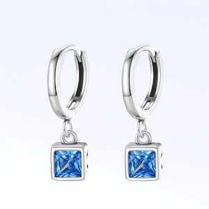 May include: A pair of silver hoop earrings with square, blue gemstone pendants. The earrings have a polished finish and a simple design. The blue gemstones are a vibrant shade of blue.