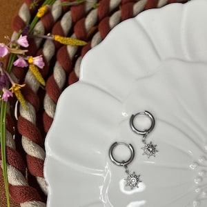 May include: Two silver hoop earrings, each with a sunburst charm, are presented on a white, scalloped plate. The background features a woven mat in red and cream, and some small flowers.