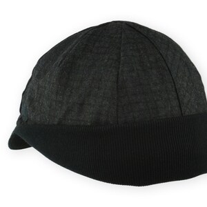 Equinox Winter Cap by Red Dots Cycling. Detail of black, double-layered, bamboo cotton ribbed ear flap in up position. 100% wool plaid fabric, charcoal grey/black plaid and purple accent. Red Dots black logo embroidered on side panel.