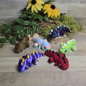 Puede incluir: Una colección de figuras de dragones coloridos hechas a mano. Los dragones son de varios colores, incluyendo marrón, morado, rojo, azul y verde, con detalles dorados o blancos. Están dispuestos sobre una superficie de madera con flores y vegetación.