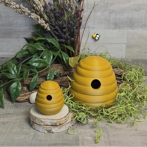 May include: Two yellow beehive-shaped decorative items. The larger one is on a bed of green moss, and the smaller one sits on a wooden slice. A small, decorative bee is in the background. The items are for home decor.