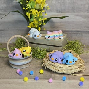 May include: A set of five colourful, hand-painted bird figurines. The birds are in various poses, including sitting, standing, and flying. The figurines are made of a smooth, ceramic-like material and have a glossy finish. The birds are decorated with bright colours, including pink, yellow, blue, and purple. The figurines are displayed on a wooden surface with a basket and a nest.