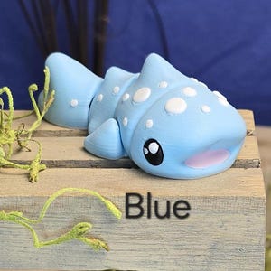May include: A light blue, 3D-printed whale shark figurine with white spots, black eyes, and a pink mouth. The figurine is displayed on a wooden surface with the word "Blue" printed on it.