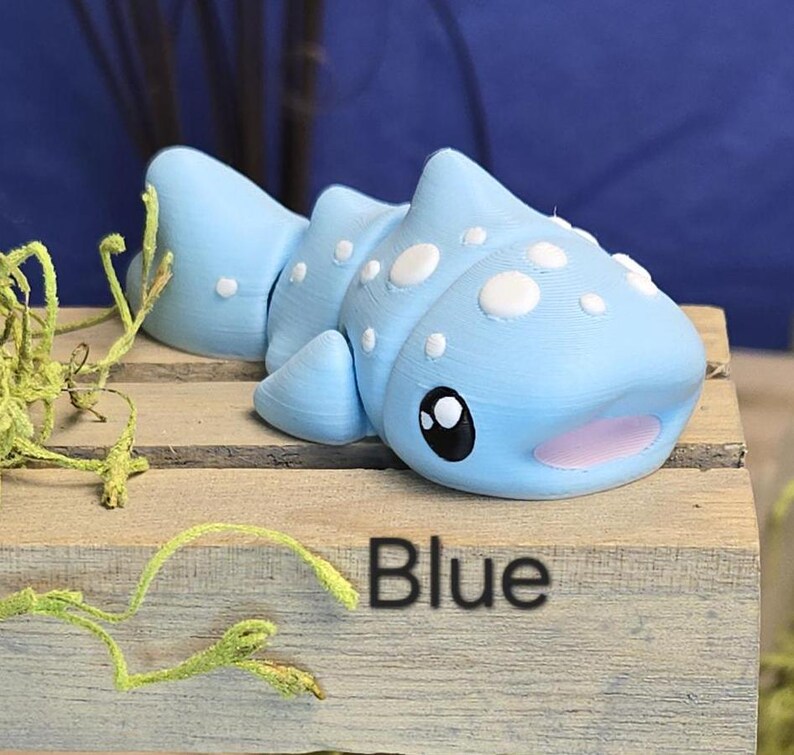 May include: A light blue, 3D-printed whale shark figurine with white spots, black eyes, and a pink mouth. The figurine is displayed on a wooden surface with the word "Blue" printed on it.
