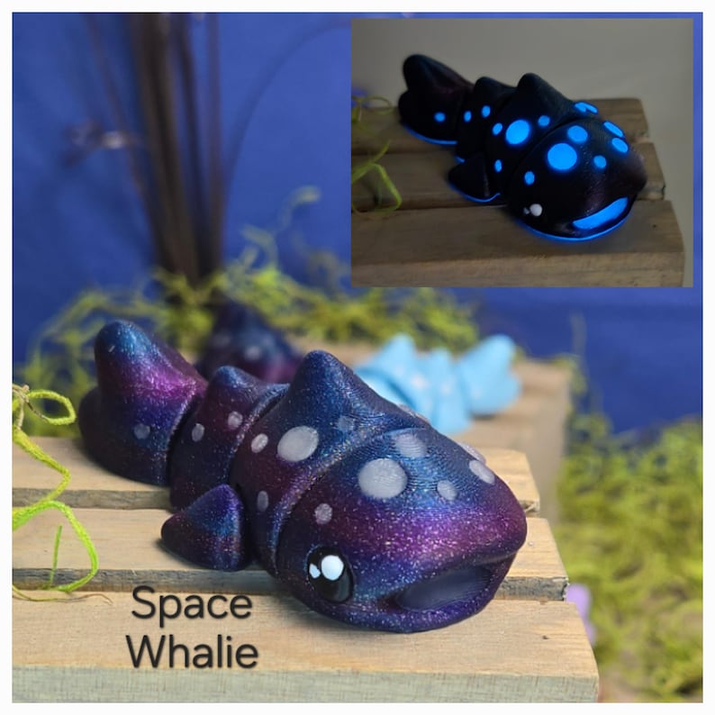 May include: A whimsical, galaxy-themed Space Whalie figurine. The sculpture features a gradient of purple, blue, and silver glitter, with white dot accents. The text "Space Whalie" is visible on the base. A smaller, glowing version is also shown.
