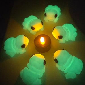 Puede incluir: Un grupo de seis figuritas verdes y negras que brillan en la oscuridad dispuestas en círculo alrededor de una pequeña vela. Las figuritas tienen forma de orugas con ojos negros y un cuerpo segmentado.