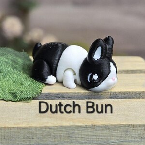 K&ouml;nnte beinhalten: Eine 3D-gedruckte Figur eines holl&auml;ndischen Kaninchens, das liegt. Das Kaninchen hat ein schwarz-wei&szlig;es Muster mit schwarzen Ohren und einem schwarzen Fleck um die Augen. Der Text "Dutch Bun" ist darunter gedruckt.