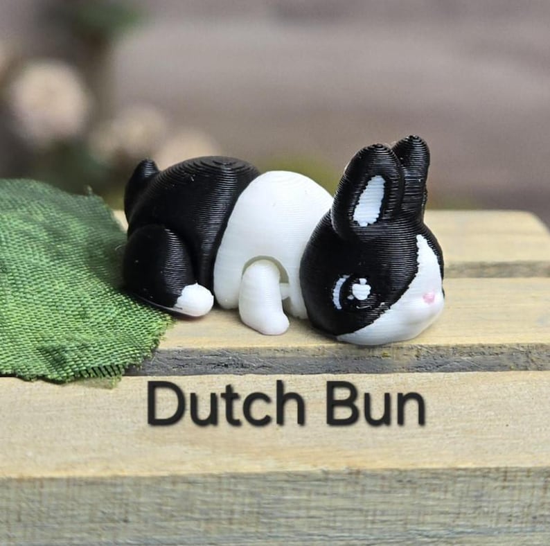 K&ouml;nnte beinhalten: Eine 3D-gedruckte Figur eines holl&auml;ndischen Kaninchens, das liegt. Das Kaninchen hat ein schwarz-wei&szlig;es Muster mit schwarzen Ohren und einem schwarzen Fleck um die Augen. Der Text "Dutch Bun" ist darunter gedruckt.