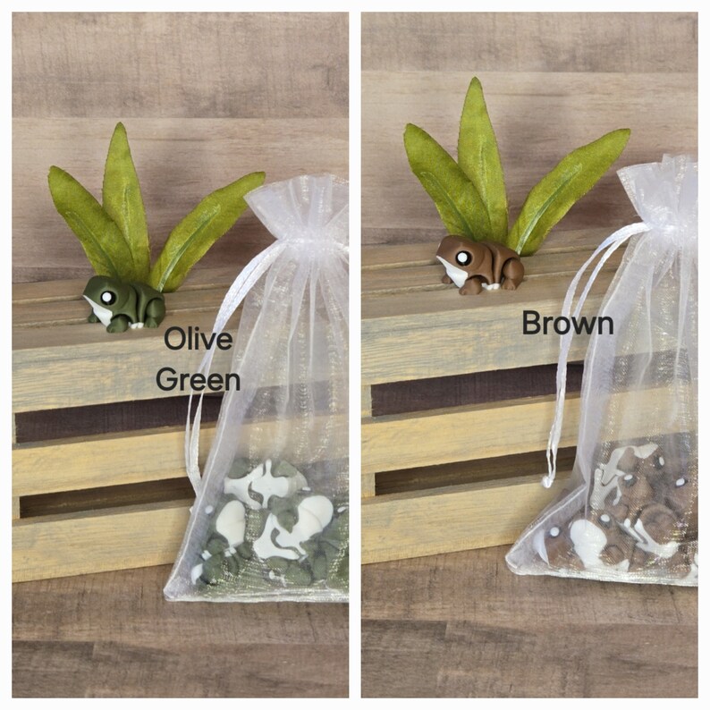 May include: Two small wooden frog figurines, one olive green and one brown, are nestled in a white tulle bag. The frogs are surrounded by green and brown leaves. The bags are sitting on a wooden crate with the words "Olive Green" and "Brown" printed below each frog.