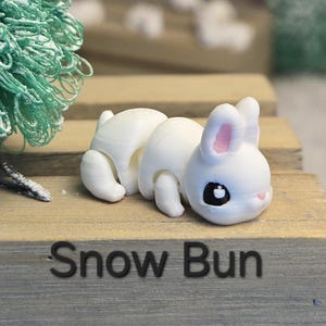 K&ouml;nnte beinhalten: Eine wei&szlig;e, 3D-gedruckte Hasenfigur mit rosa Innenohren und schwarzen Augen. Der Hase liegt auf einer Holzoberfl&auml;che. Der Text "Snow Bun" ist in Schwarz auf die Holzoberfl&auml;che gedruckt.