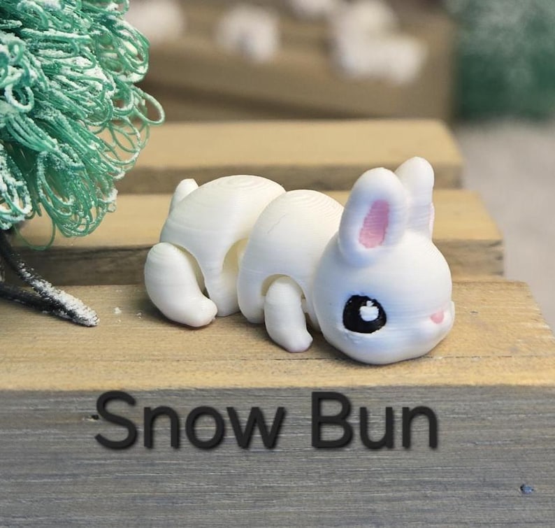 K&ouml;nnte beinhalten: Eine wei&szlig;e, 3D-gedruckte Hasenfigur mit rosa Innenohren und schwarzen Augen. Der Hase liegt auf einer Holzoberfl&auml;che. Der Text "Snow Bun" ist in Schwarz auf die Holzoberfl&auml;che gedruckt.
