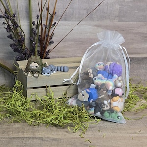 May include: A selection of miniature, brightly coloured figurines, including a grey raccoon and a brown creature, are arranged on a wooden crate. A sheer white bag filled with more figurines is also present. Green moss and purple-flowered stems complete the scene.