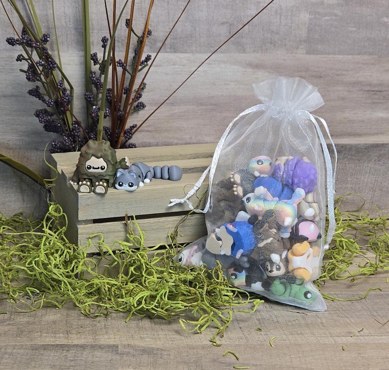 May include: A selection of miniature, brightly coloured figurines, including a grey raccoon and a brown creature, are arranged on a wooden crate. A sheer white bag filled with more figurines is also present. Green moss and purple-flowered stems complete the scene.