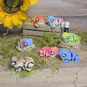 Op de afbeelding: Zeven kleine, handgeschilderde kikkerfiguurtjes met ombre kleurschema's. De kikkers zijn gerangschikt op een houten oppervlak met groen mos.