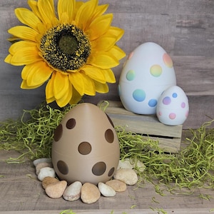 Puede incluir: Tres huevos de Pascua decorativos, uno marrón con lunares marrones, uno blanco con lunares arcoíris y uno más pequeño rosa y morado con lunares blancos. Los huevos están sobre un lecho de musgo verde con un girasol en el fondo.