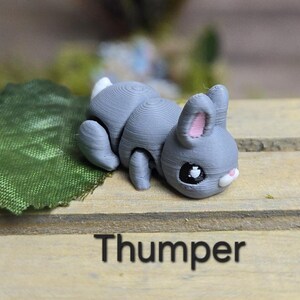 K&ouml;nnte beinhalten: Graue Hasenfigur mit wei&szlig;em Schwanz und rosa Innenohren, die auf einer Holzoberfl&auml;che liegt. Der Hase hat schwarze Augen und eine wei&szlig;e Nase. Das Wort "Thumper" ist darunter in Schwarz gedruckt.