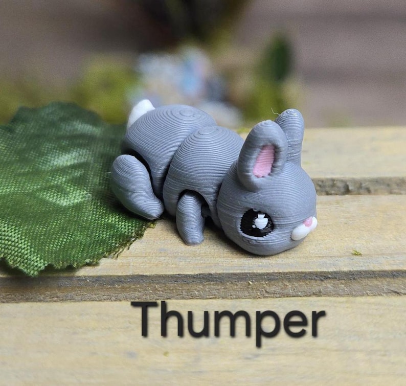 K&ouml;nnte beinhalten: Graue Hasenfigur mit wei&szlig;em Schwanz und rosa Innenohren, die auf einer Holzoberfl&auml;che liegt. Der Hase hat schwarze Augen und eine wei&szlig;e Nase. Das Wort "Thumper" ist darunter in Schwarz gedruckt.