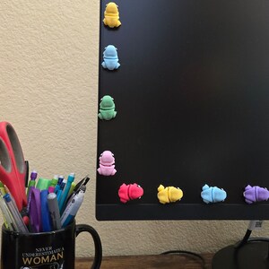 May include: A black computer monitor with a variety of colorful frog figurines attached to the edge. The monitor is on a wooden desk with a black mug, a keyboard, and two sticky notes.