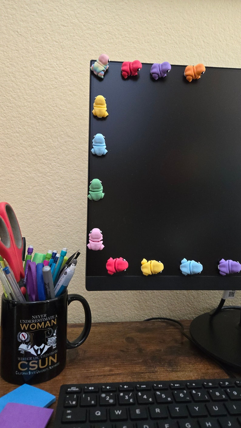 May include: A black computer monitor with a variety of colorful frog figurines attached to the edge. The monitor is on a wooden desk with a black mug, a keyboard, and two sticky notes.