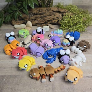 May include: A collection of colorful, handcrafted polymer clay figurines in various shapes, including frogs, scorpions, and caterpillars. The figurines are in shades of orange, pink, purple, blue, and green, with white and black accents. The collection is arranged on a wooden surface.
