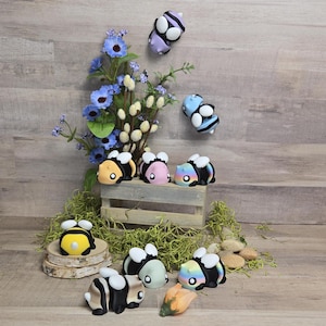 Puede incluir: Una colección de figuritas de abejas coloridas con diferentes patrones y diseños. Las abejas están hechas de un material liso y brillante y tienen alas blancas. Algunas de las abejas están sentadas en una caja de madera, mientras que otras están esparcidas sobre una superficie verde y musgosa.