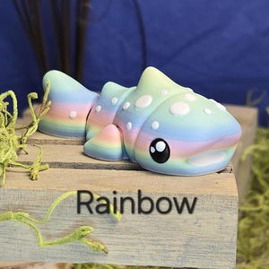 May include: A colorful, rainbow-themed sculpture of a stylized fish with white spots and large black eyes. The word "Rainbow" is printed on the wooden surface below the fish. The fish is resting on a wooden surface.
