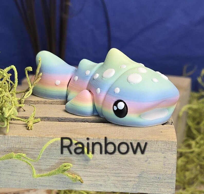 May include: A colorful, rainbow-themed sculpture of a stylized fish with white spots and large black eyes. The word "Rainbow" is printed on the wooden surface below the fish. The fish is resting on a wooden surface.