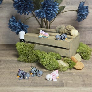 Può includere: Cinque piccole figurine di drago in ceramica dipinte a mano in vari colori, tra cui rosa, blu, verde e grigio. I draghi sono in posa in modo giocoso e fantasioso, con le ali spiegate e le code arrotolate. Le figurine sono esposte su una cassa di legno con muschio e pietre intorno.