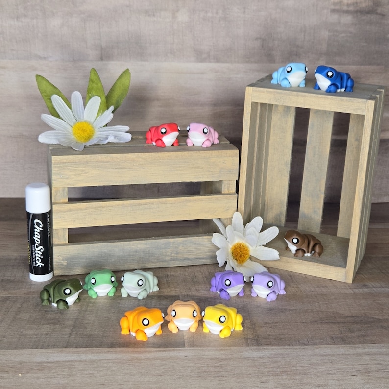 May include: A collection of colorful frog figurines, some with painted eyes, arranged on a wooden crate and a table. The frogs are in various colors, including red, pink, green, blue, purple, orange, and brown.
